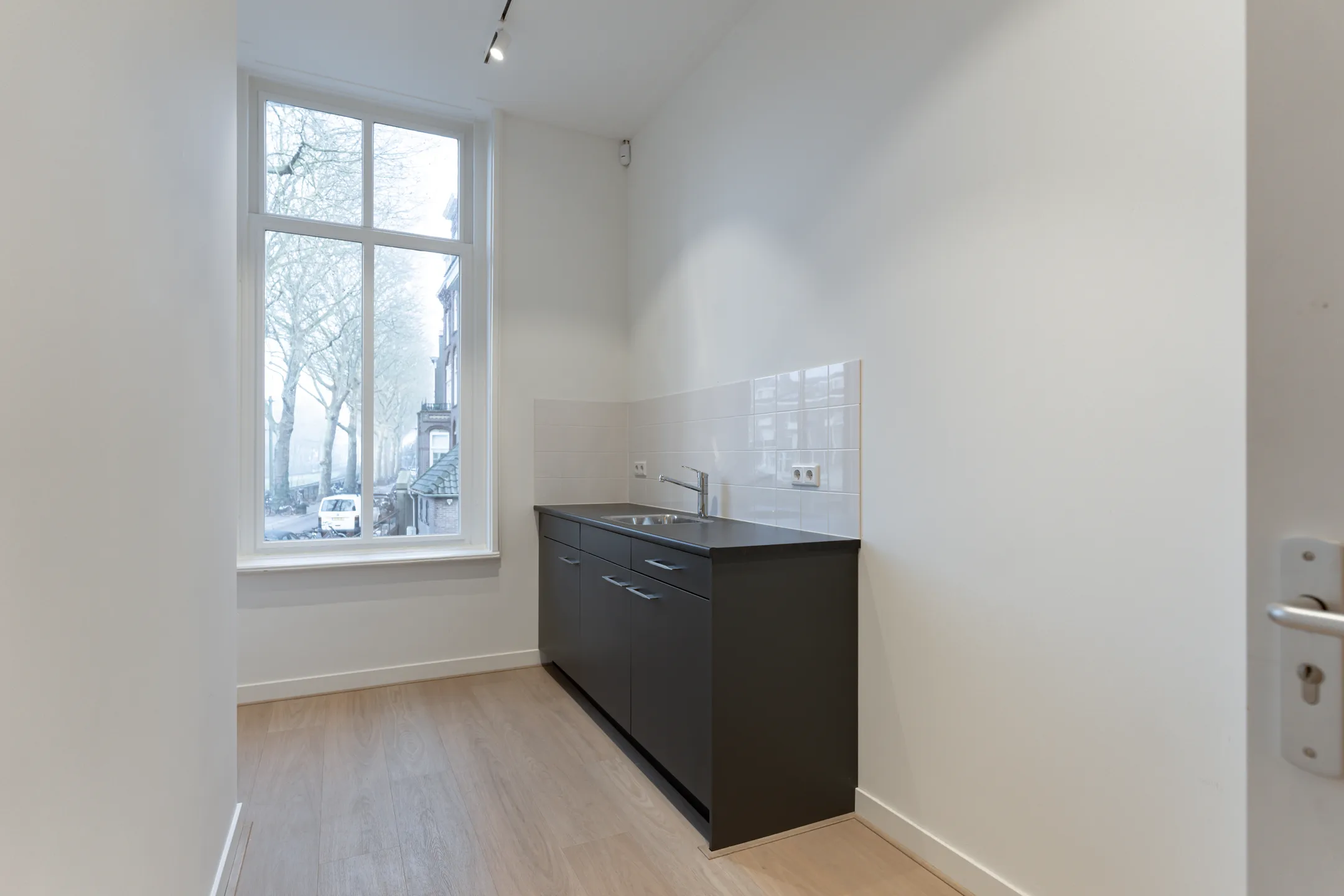 Een pantry met zwart keukenblok, spoelbak en een hoog raam met uitzicht op straat